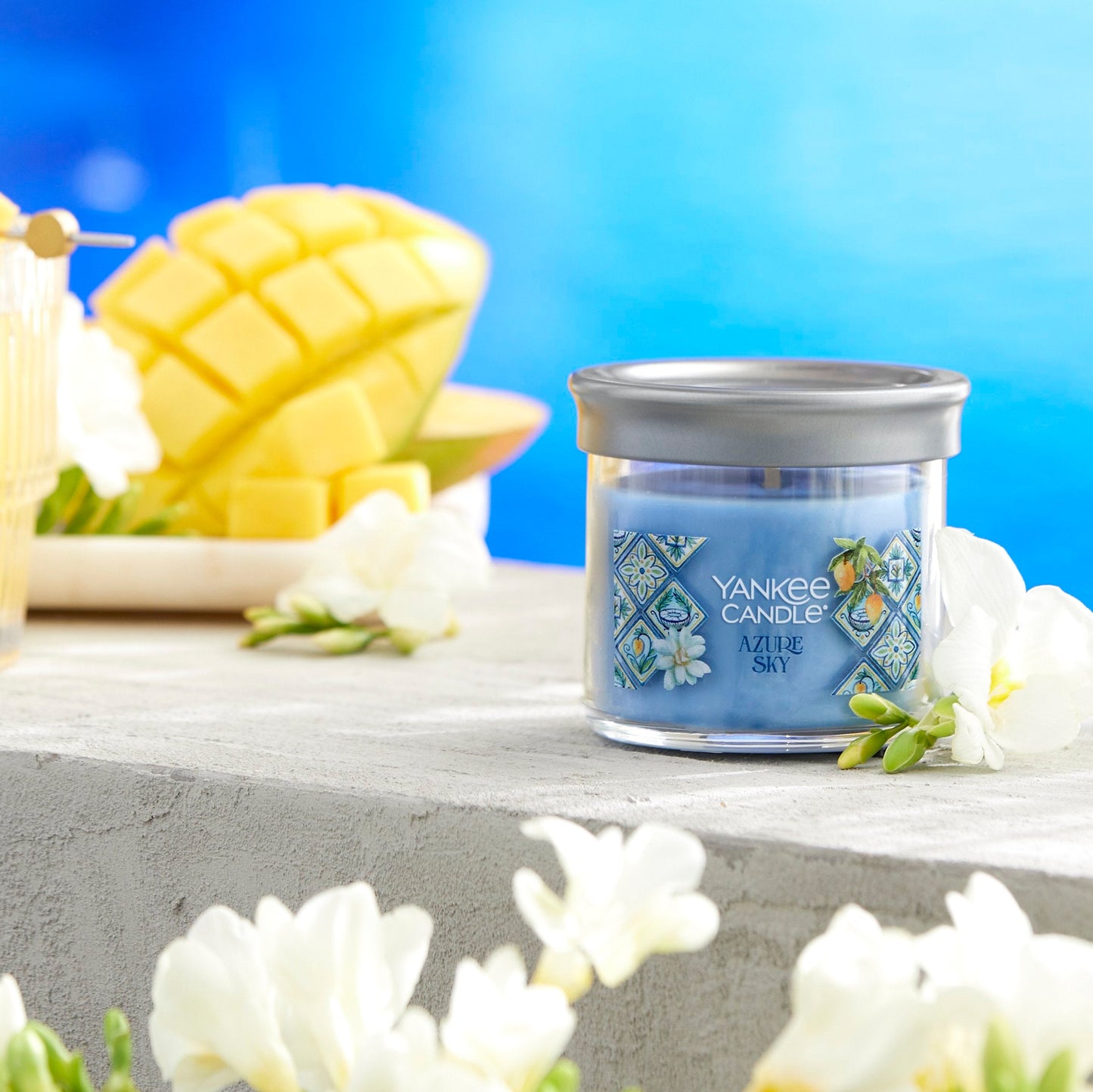 Hello, Italy! Collection Azure Sky Signature Small Tumbler Candle next to white flowers, sliced fruit, and a mixed drink